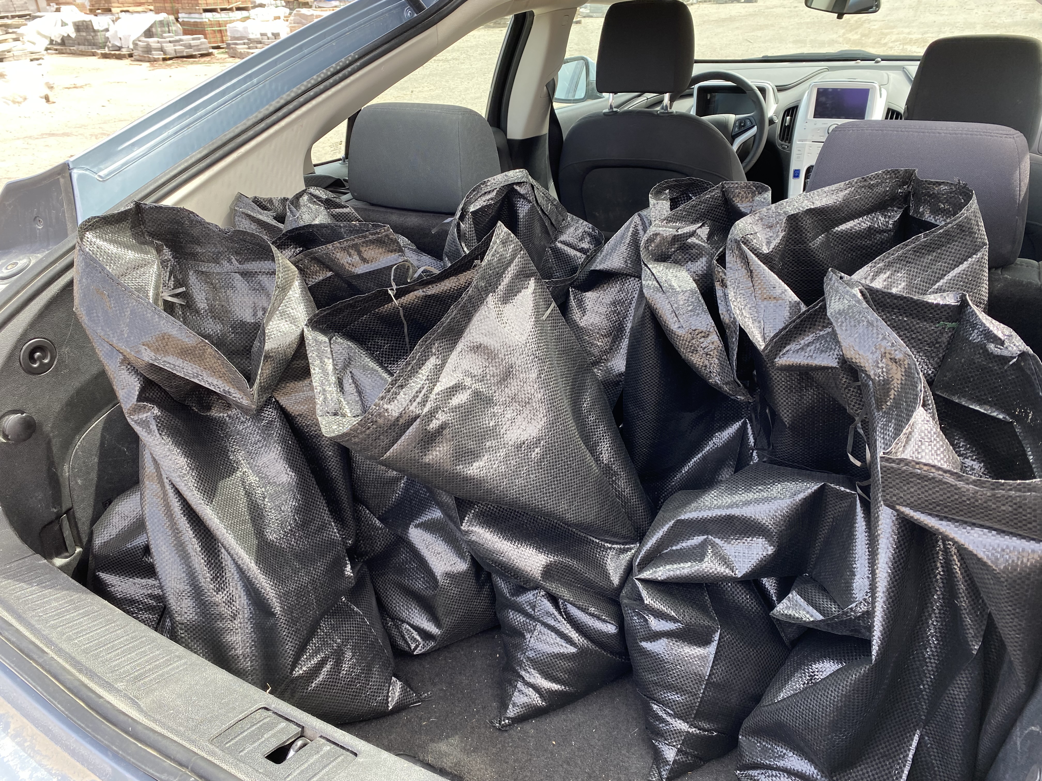 Numerous black plastic sand bags filled with sand piled in the trunk of a small car.