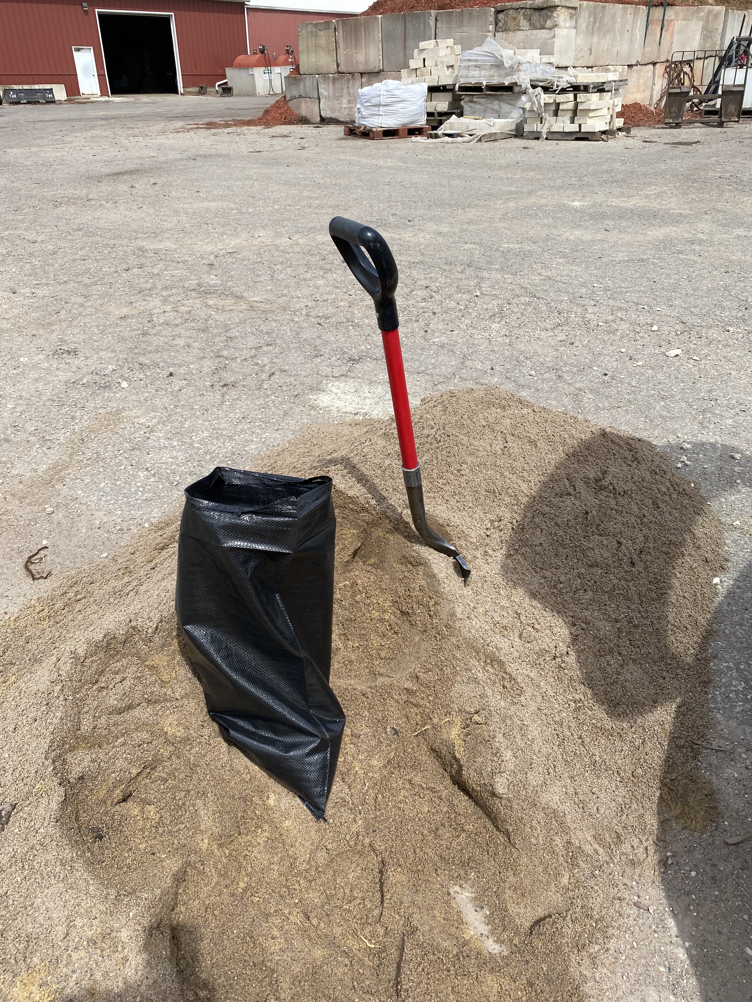Picture of shovel sticking out of a medium-sized pile of sand along with a black plastic sand bag that is being filled. The pile of sand is in the middle of a parking lot behind a nursery in an industrial looking site.